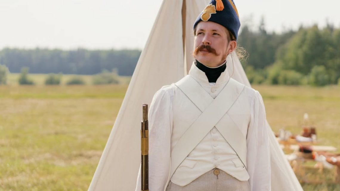 A historical reenactor in Napoleonic military uniform standing by a tent in open countryside.