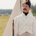 A historical reenactor in Napoleonic military uniform standing by a tent in open countryside.